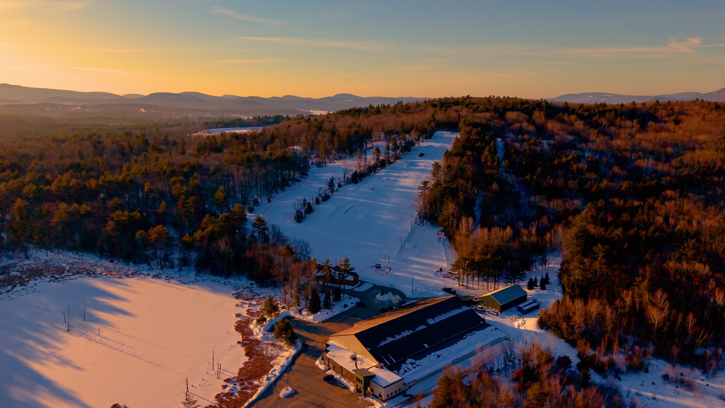 Abenaki Ski Area from the air at sunset: the lodge and parking lot in front, the main slope running up through the woods, hills and mountains beyond