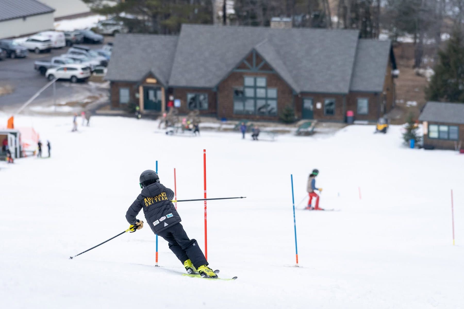 An Abenaki racer in a team jacket carving through slalom gates, with the base lodge behind