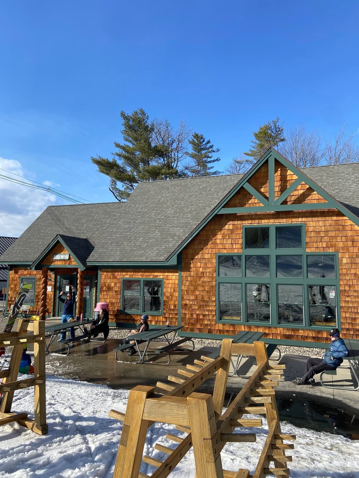 The Abenaki base lodge on a sunny afternoon, cedar shingles and green trim, with families at the picnic tables and a ski rack out front