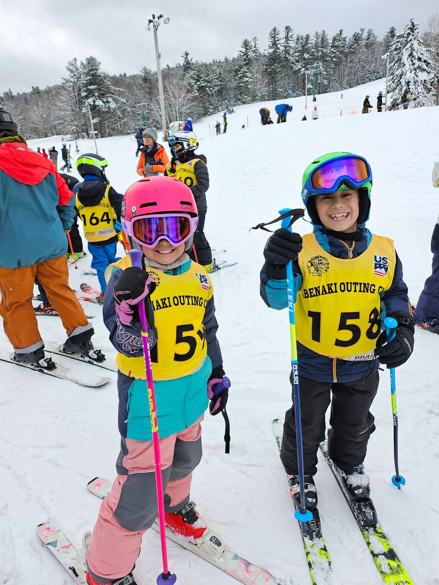Two young racers in Abenaki Outing Club bibs, poles up and grinning, at the bottom of the hill on race day