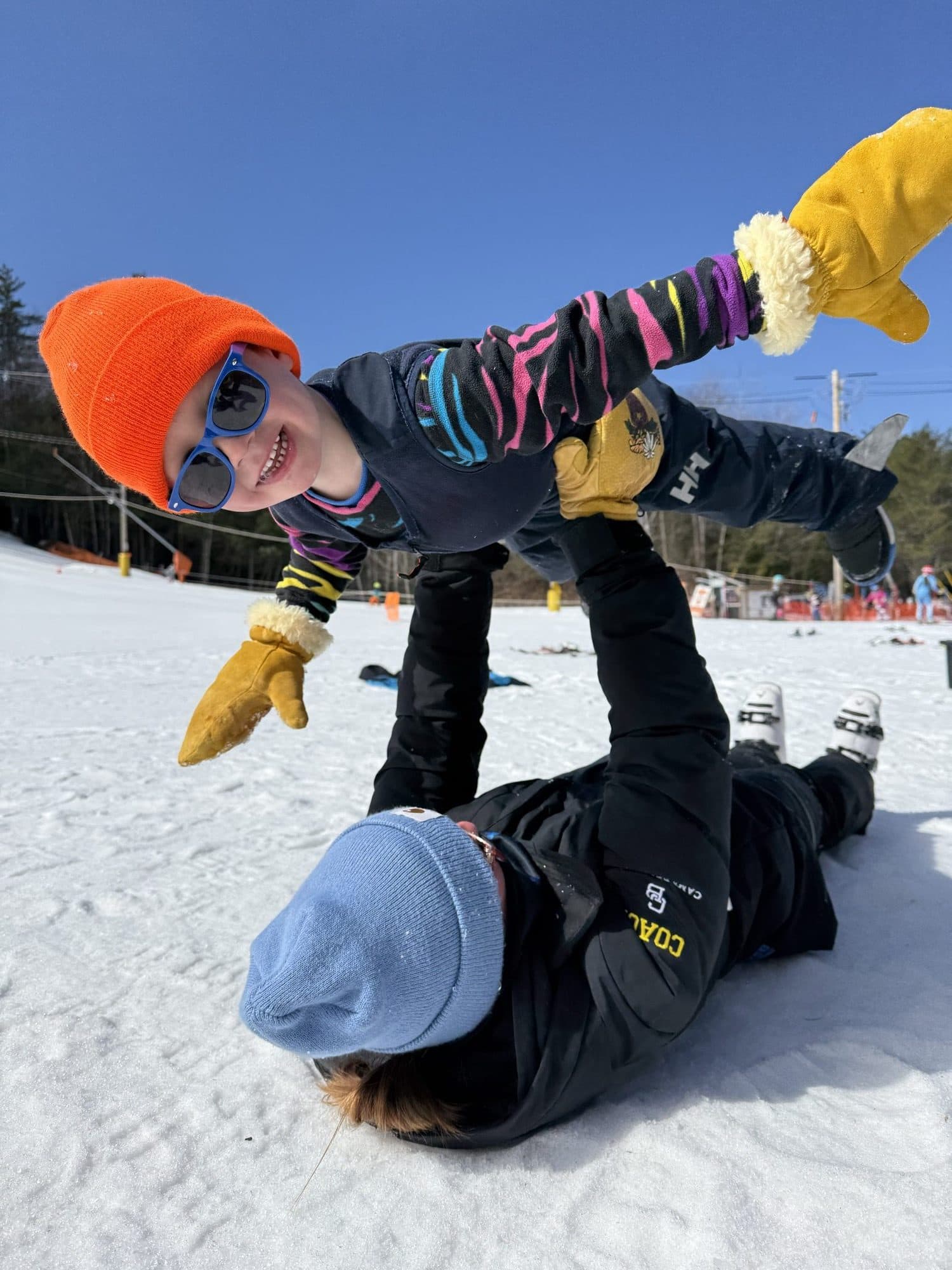 A coach on their back on the snow holding a small skier up in the air like an airplane