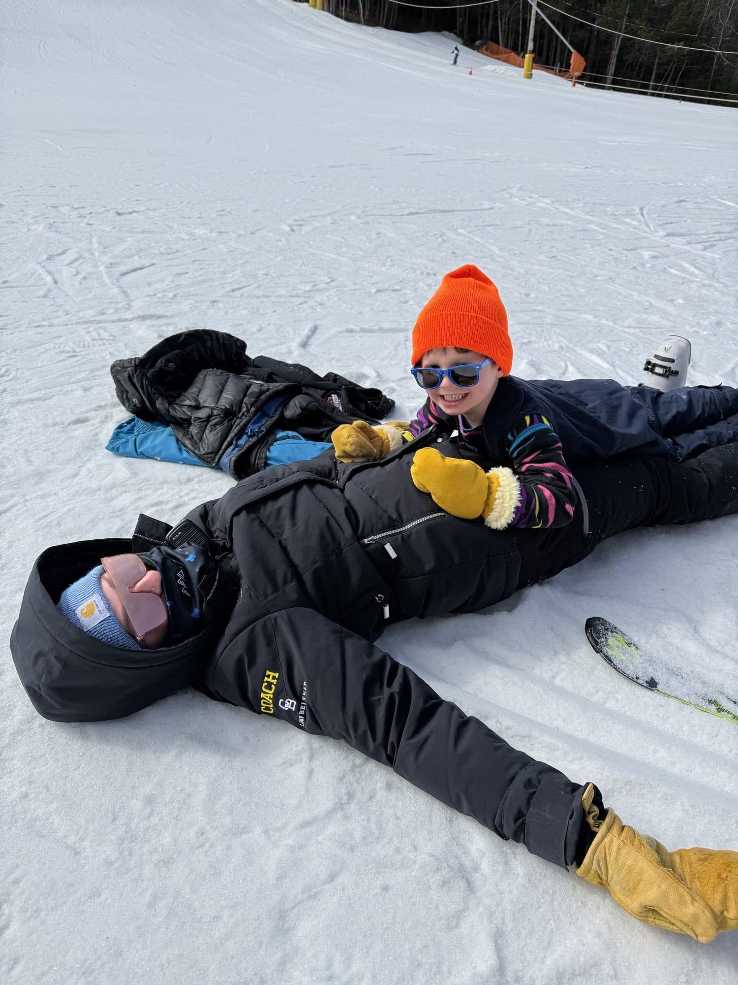 A coach lying on the snow at the bottom of the hill with a small skier in an orange hat climbing on top