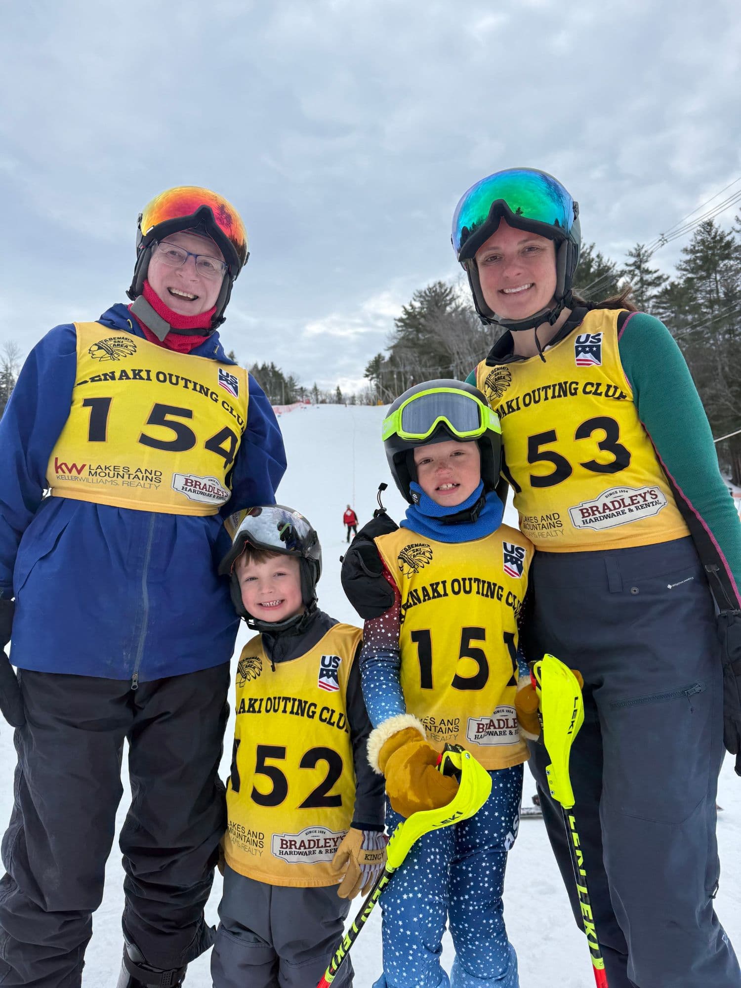 Two adults and two children in Abenaki Outing Club race bibs and helmets, standing together at the foot of the slope