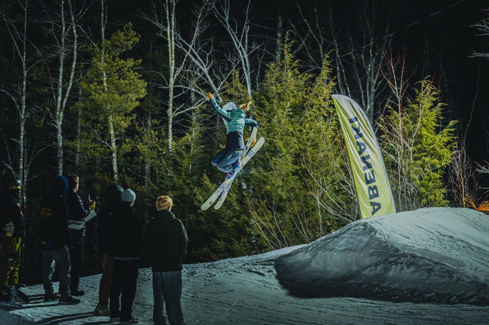 A skier grabbing their skis mid-air above the jump at night while coaches and teammates watch from the lip