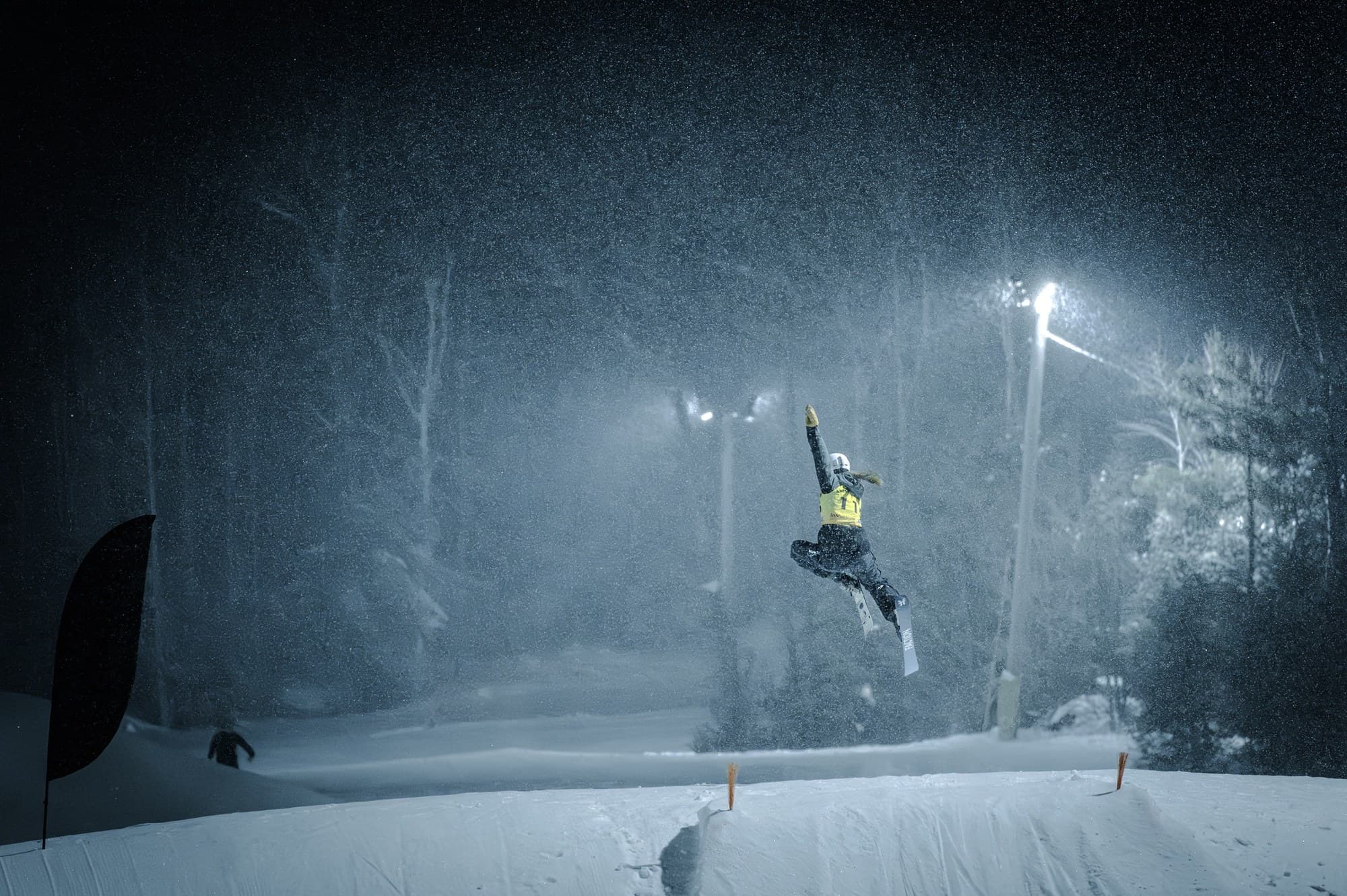 A skier in a yellow bib airborne over the jump at night, one arm up, snow streaking through the lights