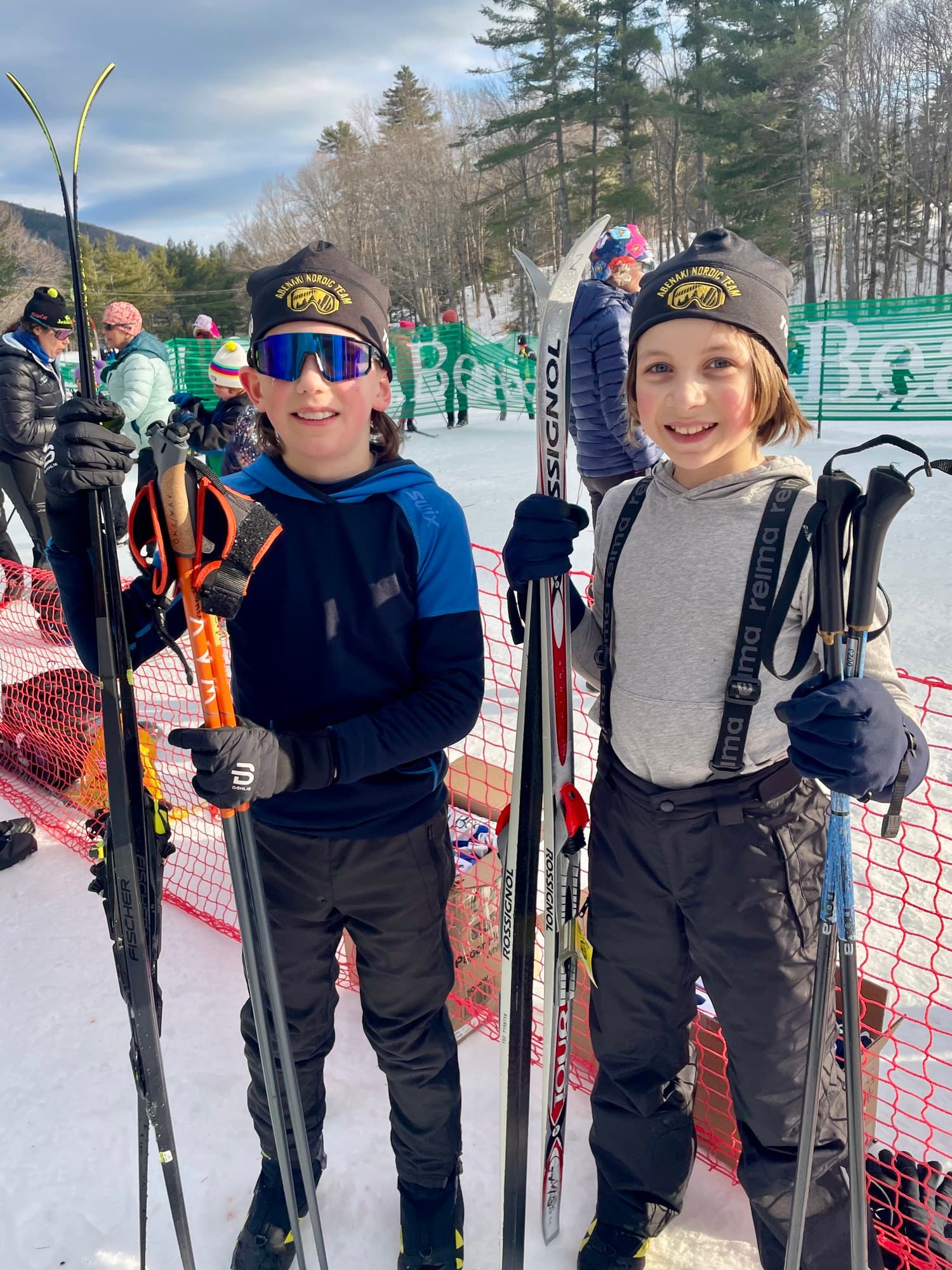 Two young skiers in Abenaki Nordic Team hats holding their skis and poles at a race