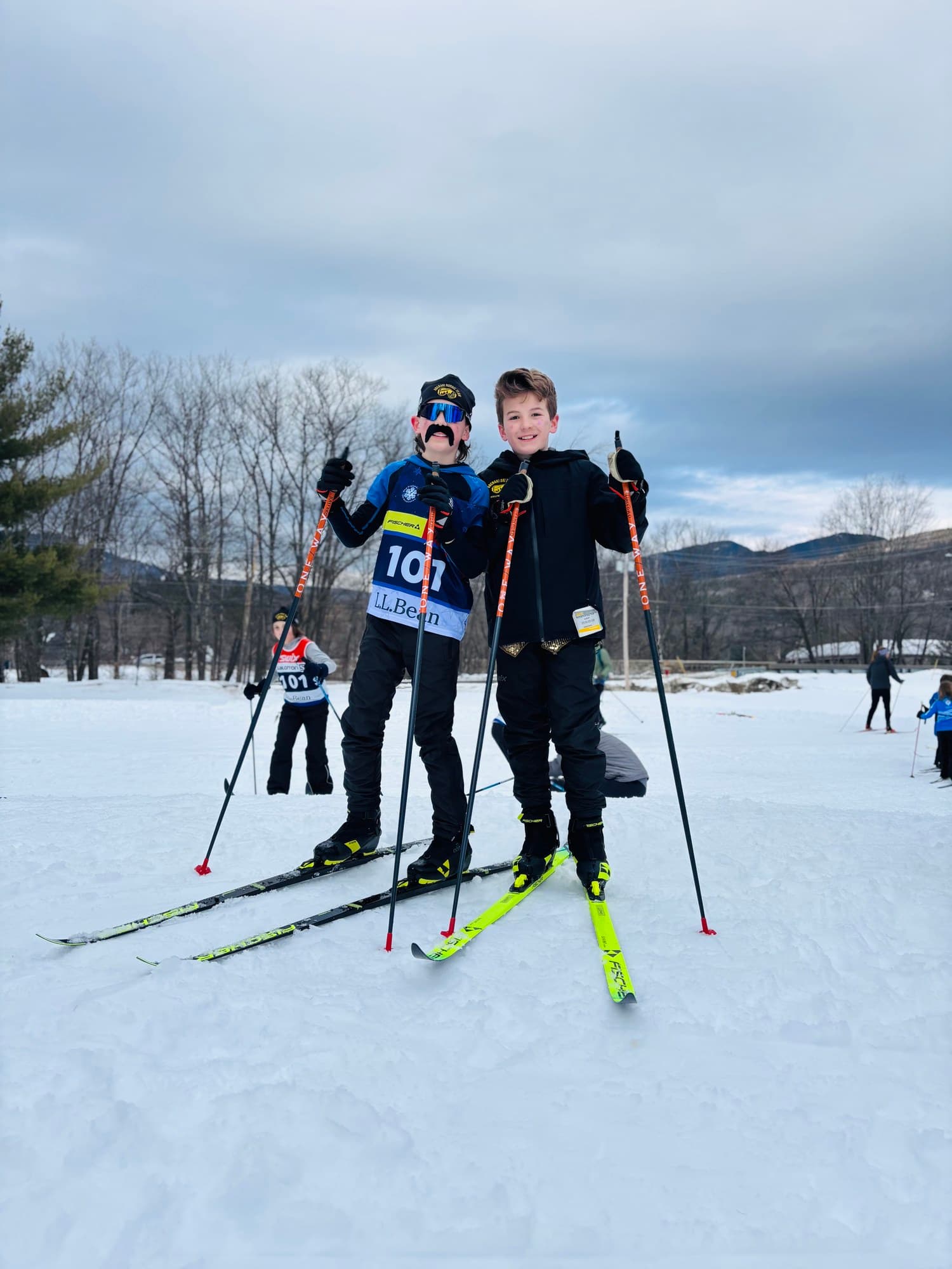 Two Nordic teammates on skis and poles at a Bill Koch race, one wearing a stick-on mustache