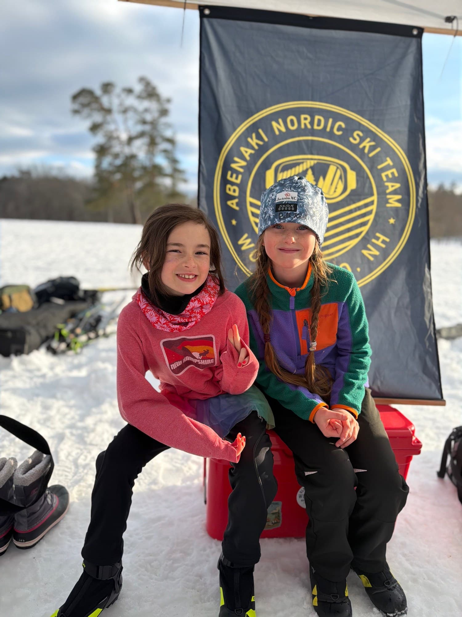 Two Nordic skiers sitting on a cooler under the Abenaki Nordic Ski Team banner