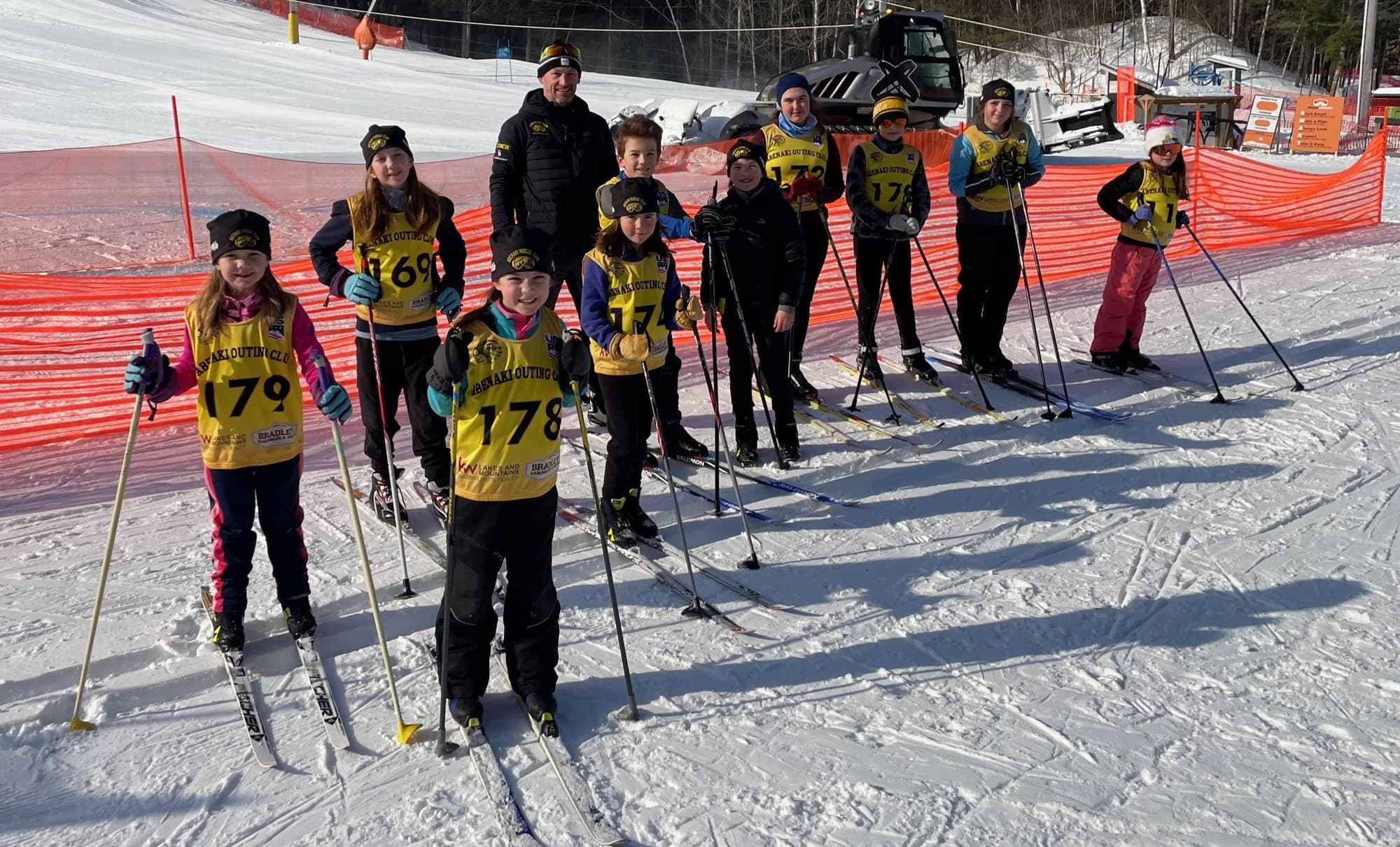 The Abenaki Nordic team in club bibs, skis and poles, lined up on the snow with their coach
