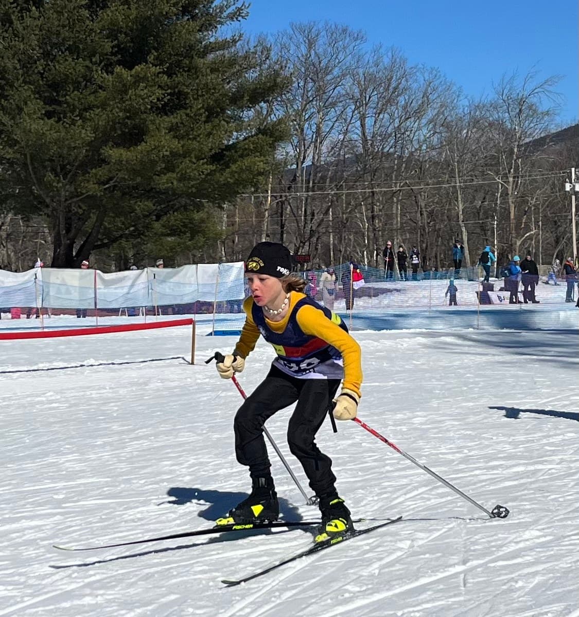 A young Abenaki Nordic skier in a yellow jacket skating hard past the course fencing at a race, spectators along the far side