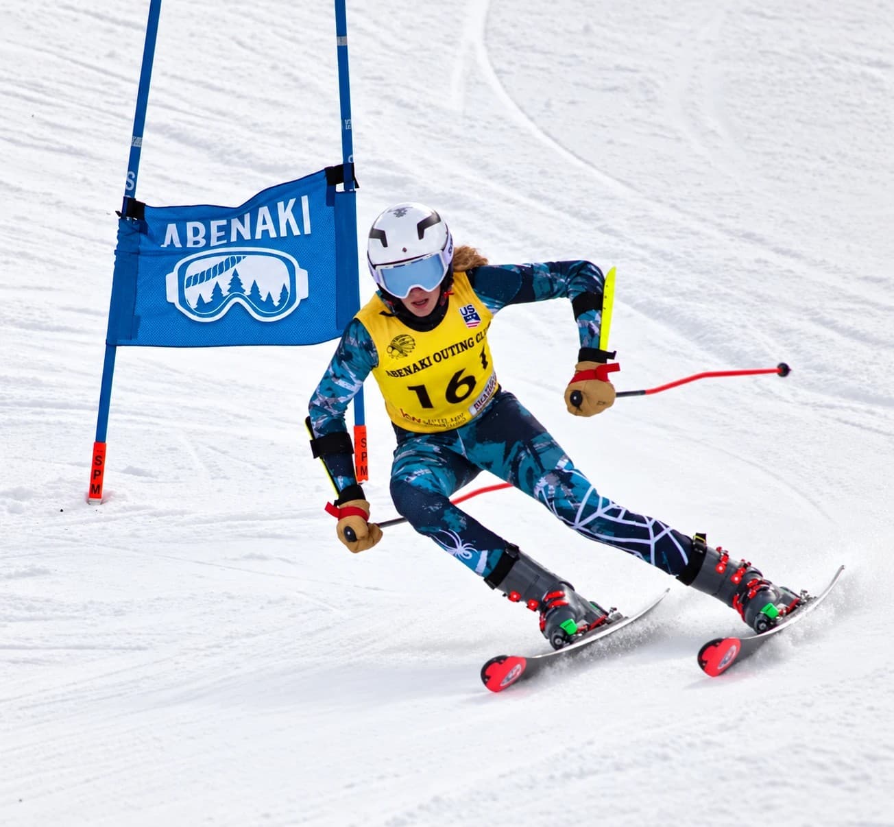 A U14 racer in Abenaki Outing Club bib 16 carving a giant slalom turn past a gate with the Abenaki goggle panel
