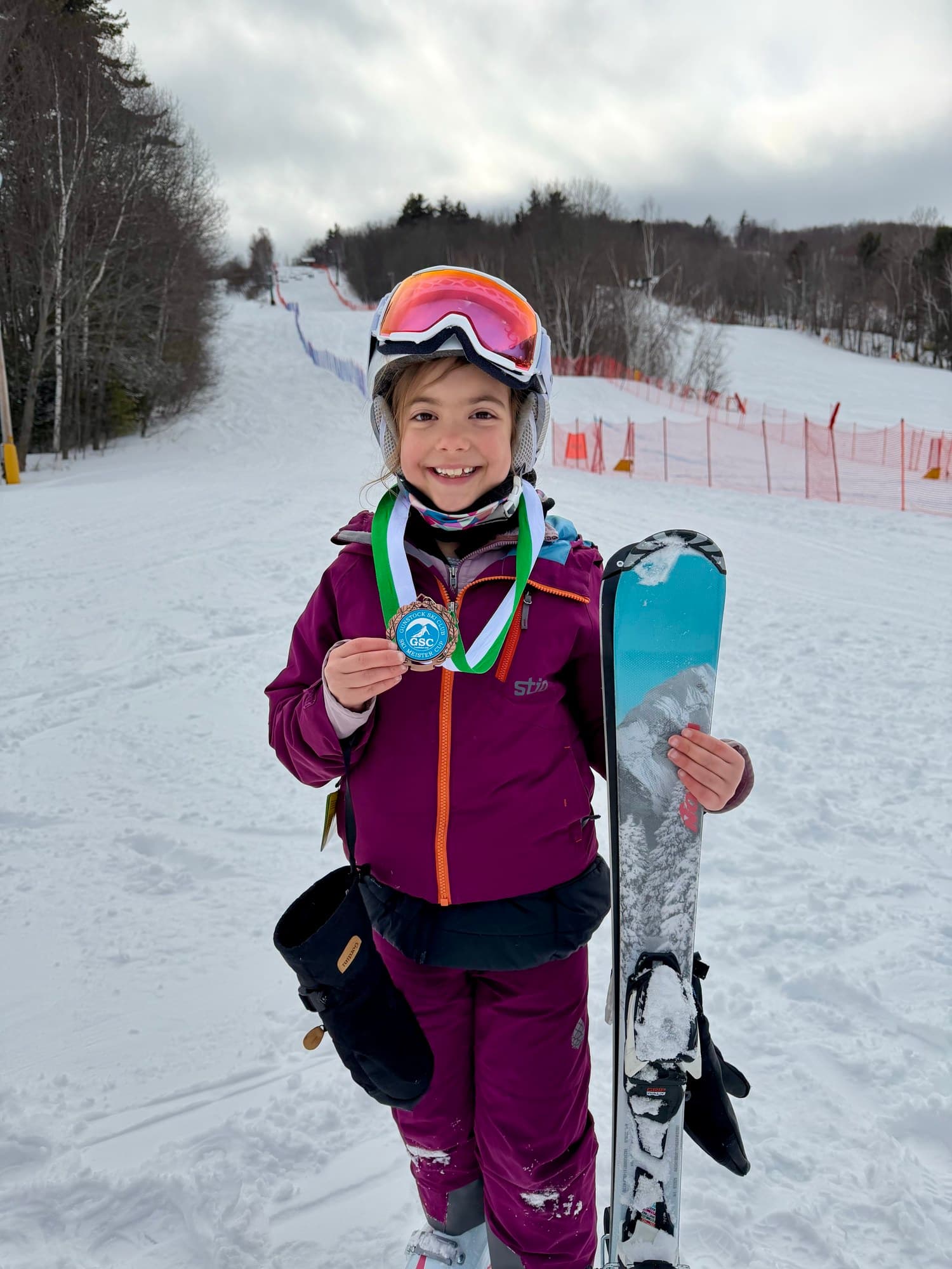 A U8 racer holding up a Gunstock Ski Club medal beside one ski, the course behind