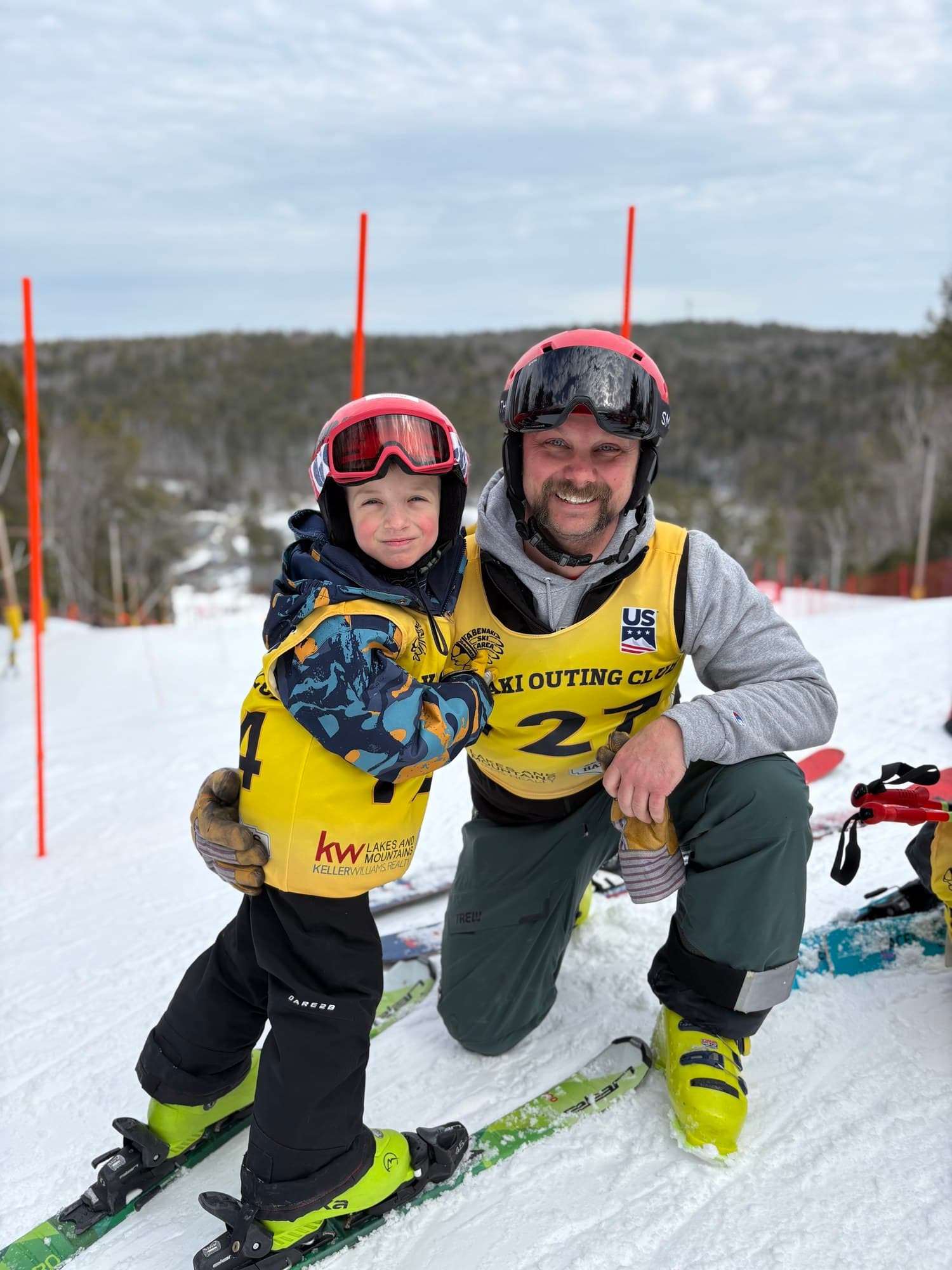 A parent in a volunteer bib kneeling beside a young racer at the top of the course