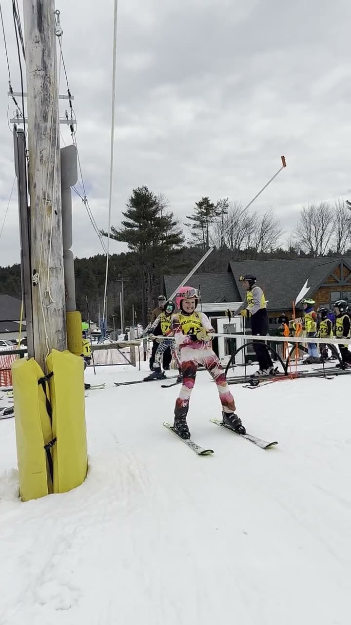 Two young racers riding the rope tow up past the tower, the tow line running up the slope at dusk, and a group of small skiers coming down the main slope together